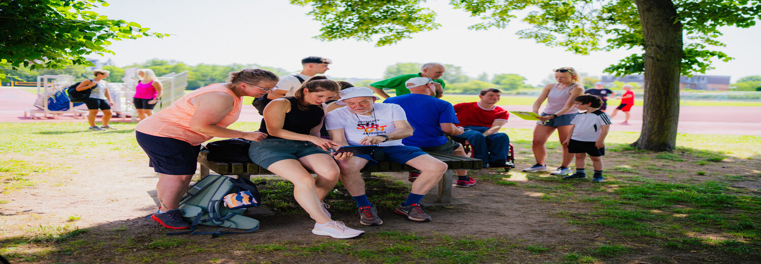 Eine Gruppe Menschen unterschiedlichen Alters sitzen auf einer Bank oder stehen um die Bank herum. Eine Person hat ein Tablet auf dem Schoß, in dem Werte der Prüfung zum Deutschen Sportabzeichen eingetragen werden.